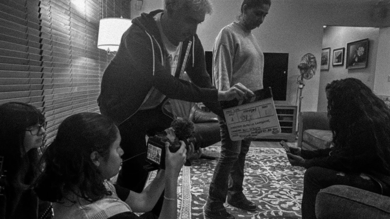 A student crew setting up a shot in a living room, one holding the slate and another on camera